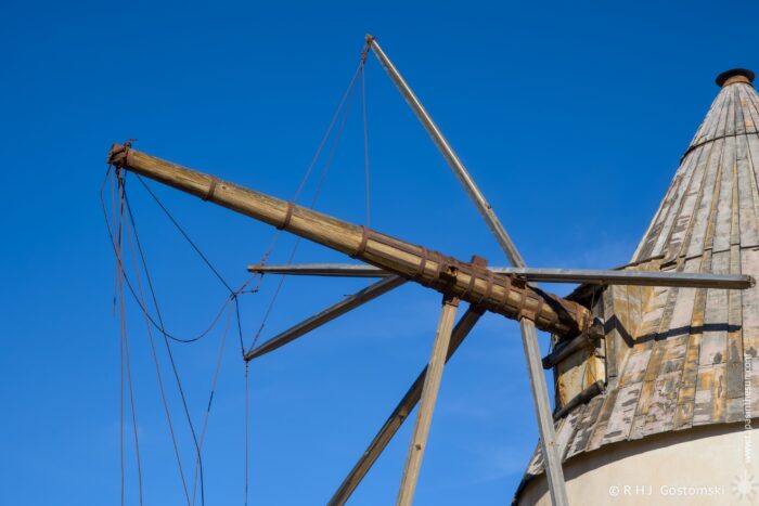 Close-up of a wooden windmill structure featuring a long arm with cables against a clear blue sky. The weathered rooftop showcases traditional design elements typical of historic mills.