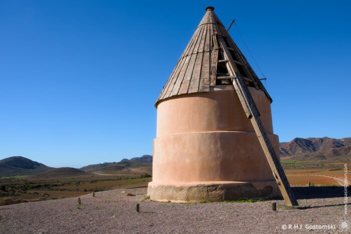 The Collado de los Genoveses windmill with a wooden roof, situated on a gravel landscape. The mill stands against a clear blue sky, surrounded by rolling hills and distant mountains, showcasing a scenic rural setting.