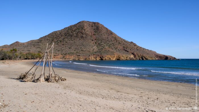 A tranquil Genoveses beach featuring a sandy shore, gentle waves lapping at the coast, and a prominent mountain in the background under a clear blue sky. A structure made of dead agave plants is placed on the beach, adding interest to the landscape.