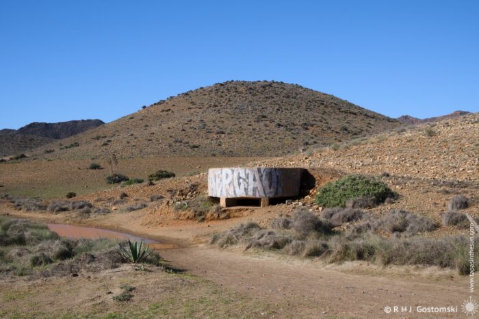 A concrete bunker with graffiti sits in a rocky, arid landscape, surrounded by sparse vegetation and rolling hills under a clear blue sky. A dirt path leads toward the structure, highlighting the natural terrain.