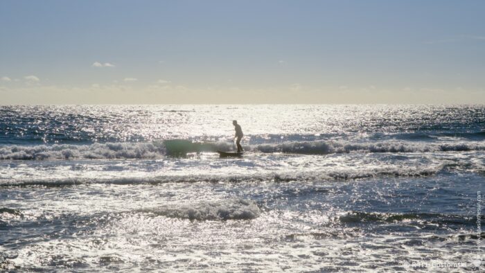 A silhouette of a person on a surfboard amongst shimmering waves, with bright reflections on the water's surface under a clear sky. The scene evokes tranquility and the beauty of nature.
