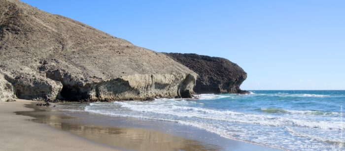 A sandy Media Luna beach with a clear blue sky. Waves gently lap against the shore, while a prominent rock formation juts out towards the sea. The landscape features sandy areas and rugged cliffs, creating a serene coastal scene.