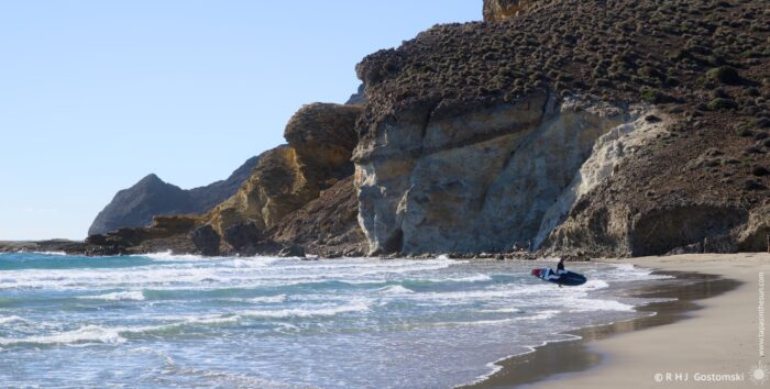 Media Luna Beach with gentle waves lapping at the shore, with rugged cliffs in the background under a clear blue sky. A surfer walks ashore across the wet sand, surrounded by a peaceful coastal landscape.