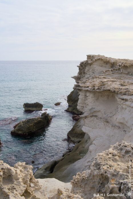 A scenic view of rugged coastal cliffs overlooking calm waters, with rocky formations in the foreground and a serene horizon under a cloudy sky.
