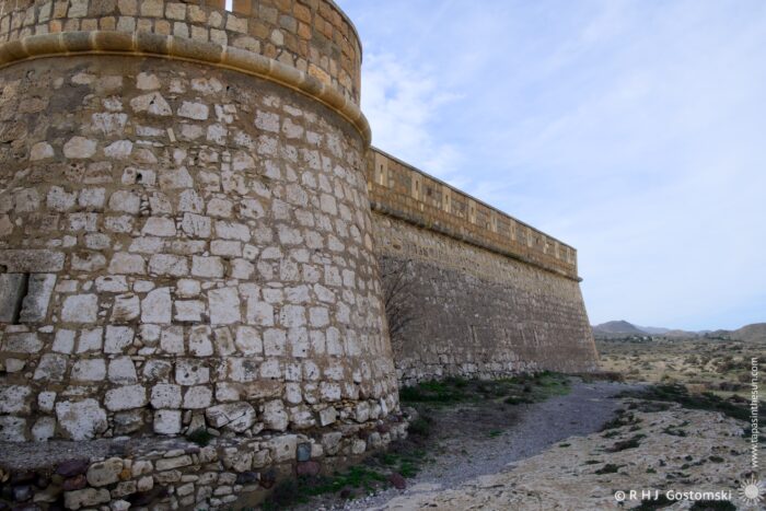 Castillo San Felipe with a round tower and a long wall, set against a backdrop of a clear blue sky and rocky landscape. The structure features weathered textures and earthy tones, highlighting its age and architectural significance.