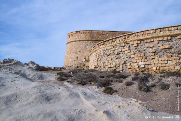 Castillo San Felipe rises on a sandy hillside, featuring rounded towers and weathered walls. The sky above is slightly cloudy, enhancing the ancient architectural beauty against a natural landscape.