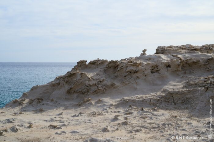 Duna F&oacute;sil de los Escullos shoreline with dramatic cliffs overlooking a calm sea under a cloudy sky. The landscape features rugged terrain and sandy areas, creating a serene coastal scene.