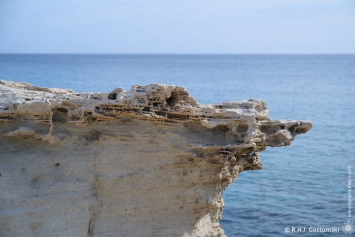 A close-up view of a weathered rock formation on Duna F&oacute;sil de los Escullos jutting out over calm sea water, with textured surfaces and eroded edges, under a clear sky.