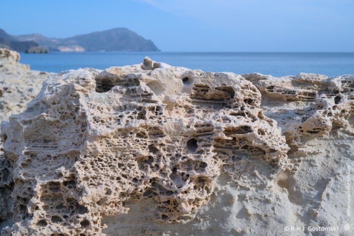 A close-up view of a textured, porous rock formation on Duna F&oacute;sil de los Escullos, with a serene ocean and distant mountains in the background under a clear blue sky.