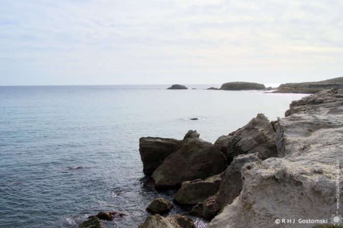 A serene Los Escullos coastal landscape featuring rocky shorelines extending into a calm sea under a cloudy sky, with distant islands visible on the horizon. The soft waves gently lap against the rocks, creating a tranquil atmosphere.