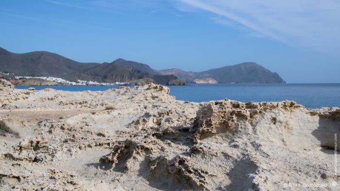 A rocky los Escullos  coastline with a sandy beach leads to calm blue waters, surrounded by mountains under a clear sky. In the distance, white buildings are visible along the shore.
