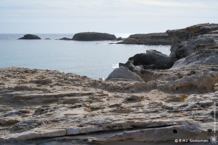 Duna F&oacute;sil de los Escullos coastline with calm waters under a cloudy sky. The scene features rugged cliffs and distant islands, creating a serene seaside landscape.