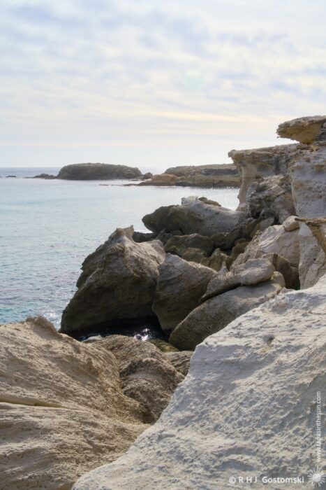 A serene Duna F&oacute;sil de los Escullos coastal scene featuring rugged gray rocks along the shoreline, gently lapping waves, and a calm sea under a cloudy sky. The landscape showcases natural formations and textures, evoking a peaceful atmosphere.