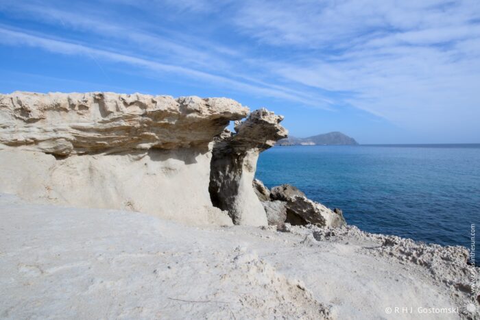 Duna F&oacute;sil de los Escullos under a blue sky, featuring a unique stone formation jutting out over the calm, clear waters of the ocean, with distant hills visible on the horizon.