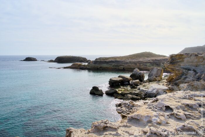 A tranquil coastal scene featuring rocky shores and calm waters under a subtle overcast sky. The landscape includes distant hills and scattered boulders, creating a serene and natural atmosphere.