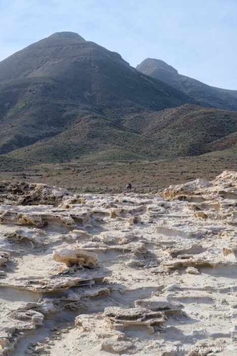 A rugged landscape featuring dry, rocky terrain with unusual formations in the foreground and distant mountains under a clear sky. A small figure is visible, adding scale to the vast, arid environment.