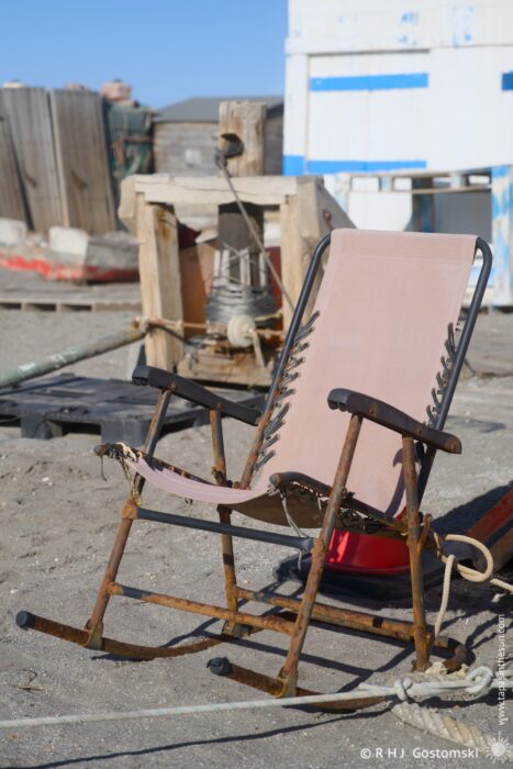 A weathered rocking chair on a beach with scattered wooden structures and equipment in the background, set against a clear blue sky.