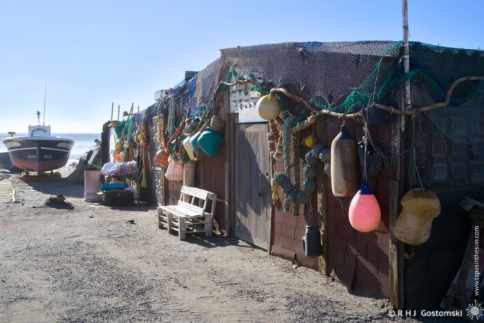 A seaside fisherman&rsquo;s shed adorned with colorful fishing buoys and nets, alongside a small boat on a sandy shore. Clear blue skies create a serene coastal atmosphere.