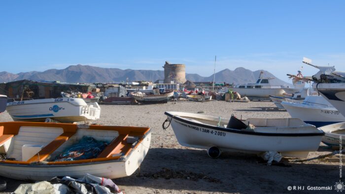 A sunny beach scene featuring several boats on sandy shores, with a distant stone tower and mountains in the background. The boats are parked closely together, creating a picturesque harbor atmosphere.