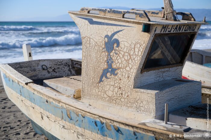 An aged fishing boat sits on the beach, showcasing a cracked white and blue paint job. The boat's stern features intricate designs, with the water and distant mountains in the background, creating a scenic coastal atmosphere.