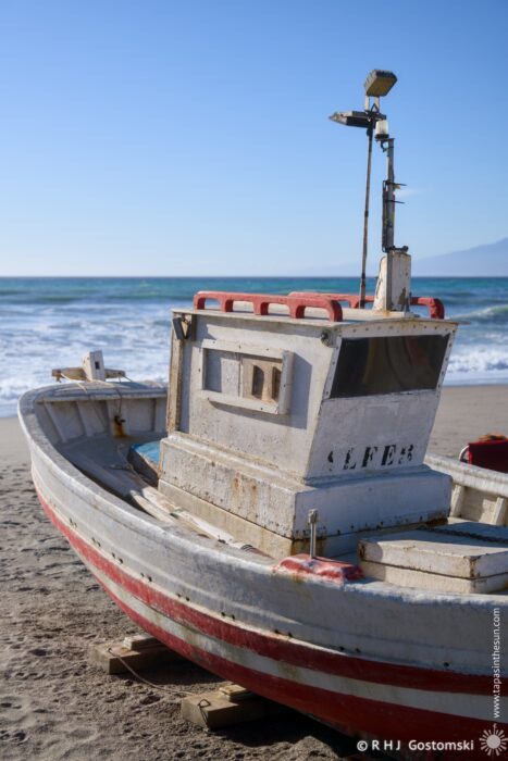 A weathered fishing boat with red and white stripes sits on a sandy beach, facing the ocean under a clear blue sky. Waves gently lap at the shore, and distant mountains can be seen on the horizon.