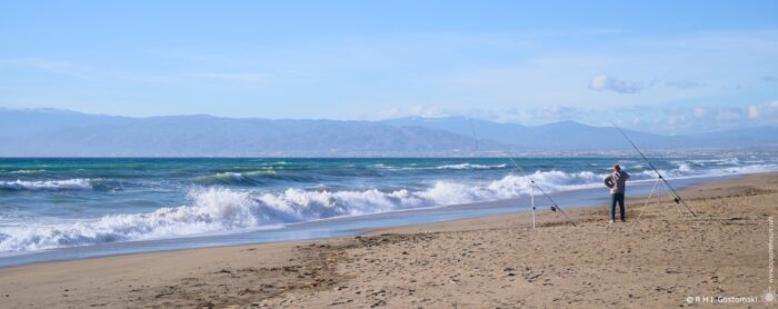 A person fishing on a sandy beach, with gentle waves lapping the shore under a clear blue sky and distant mountains in the background.