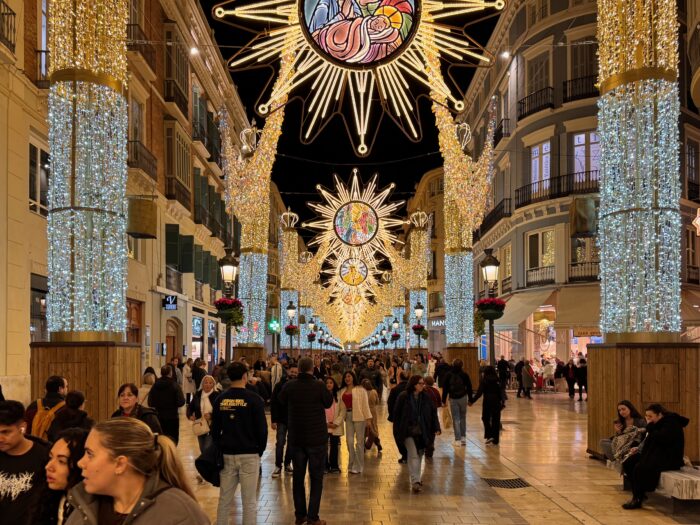 Calle Larios in Malaga illuminated with festive lights and decorations, bustling with people during nighttime. Colorful chandeliers and hanging ornaments create a joyful atmosphere, capturing the essence of a holiday celebration.
