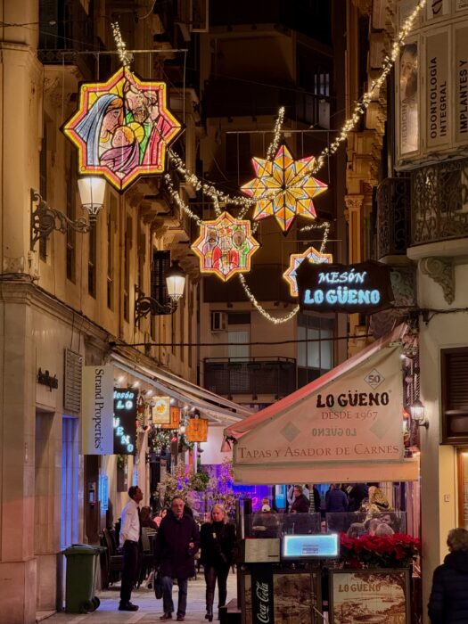A vibrant street scene illuminated by colorful Christmas lights and lanterns, featuring a restaurant sign for "Mes&oacute;n Lo G&uuml;eno" and pedestrians enjoying the festive ambiance in a bustling alleyway.
