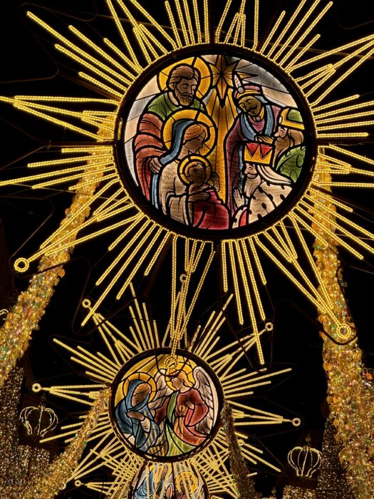 A vibrant display of illuminated stained glass decorations depicting nativity scenes, featuring figures of Mary, Joseph, and the baby Jesus, framed by radiant golden sunburst designs against a dark background.