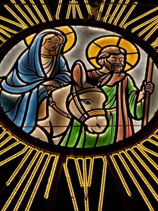 A stained glass depiction of Mary and Joseph, with Mary riding a donkey. They are surrounded by bright golden rays, symbolizing their journey. Mary is in a blue cloak, while Joseph is in green, holding a staff, conveying a sense of reverence and warmth.