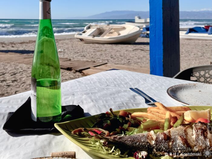 The view of Cabo de Gata beach from the Chiringuito Restaurante Chiri-Bus. In the foreground is a plate of seafood and a bottle of wine on a table.