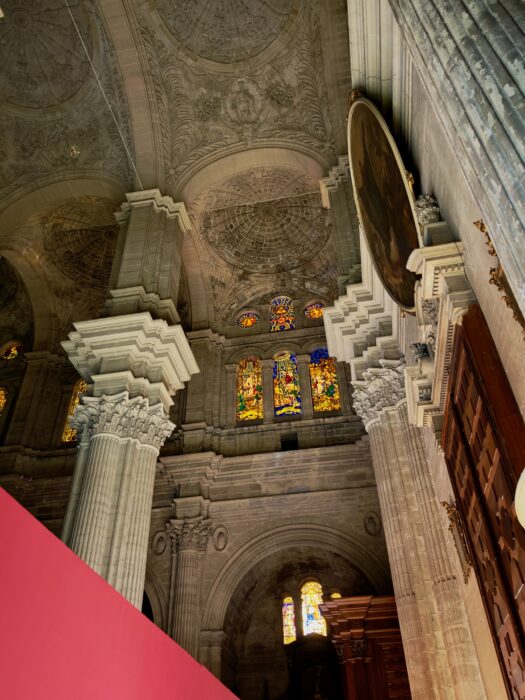Interior of Malaga Cathedral featuring intricately designed high ceilings, ornate columns, and colourful stained glass windows.