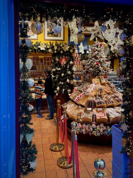 A festive shop entrance decorated with holiday ornaments features a large display of sweets and treats, surrounded by Christmas decorations, a decorated tree, and visitors admiring the offerings.