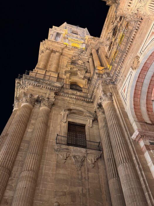 A low-angle view of a Malaga Cathedral at night, showcasing intricate architectural details, tall columns, and a prominent bell tower illuminated against the dark sky.