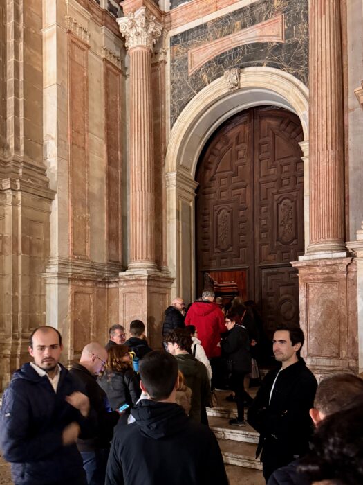A line of people waiting to enter a grand doorway with intricate designs, surrounded by elegant stone columns and rich architectural details.