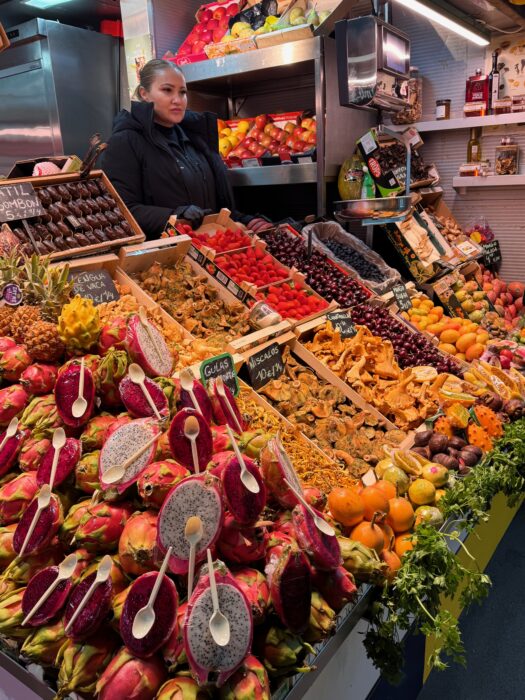 A vibrant market stall displays a colorful array of fresh fruits, mushrooms, and nuts. A woman stands behind the counter, showcasing dragon fruit, pineapples, and strawberries among other produce. The bustling atmosphere reflects a lively shopping experience.