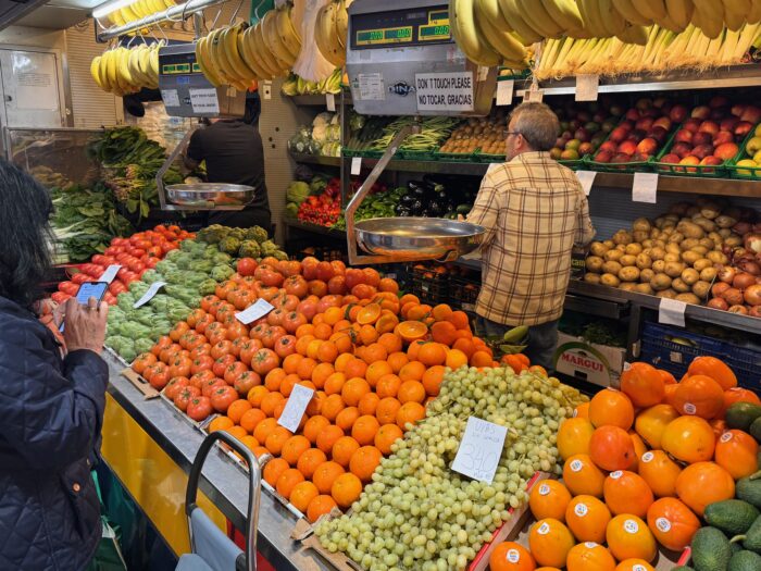 A vibrant market display featuring an array of fresh fruits and vegetables, including oranges, tomatoes, avocados, and bananas. Shoppers browse the colorful produce, creating a lively and inviting atmosphere.