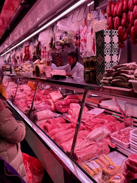 A bustling butcher&rsquo;s market stall displaying a variety of fresh meats, sausages hanging above, and a vendor in a white apron serving customers. The shop is well-organised with different cuts of meat on display, highlighting vibrant colours and textures.