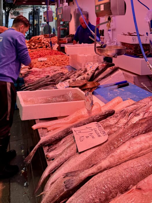 A bustling fish market scene with fish displayed on ice, a vendor preparing seafood, and customers nearby. The area is vibrant, with an array of fresh fish varieties and colorful seafood around.