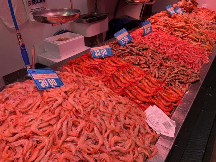 A seafood market display showcases a variety of shrimp, neatly piled in vibrant colours. Price tags in blue are visible, indicating different types and sizes, creating an appealing and fresh atmosphere typical of a fish market.