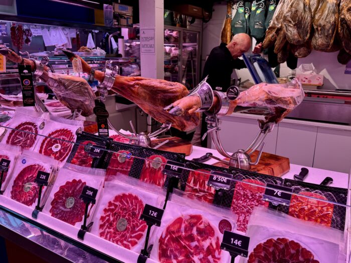 A vibrant display of various cured meats in a market, with whole jamons and neatly arranged slices in packaging. A vendor is preparing products behind the counter, showcasing a variety of prices and options for shoppers.