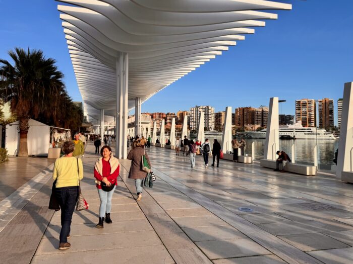 A bustling promenade lined with modern architecture, where people stroll along the waterfront in the Poprt oif Malaga. The scene features palm trees and city buildings in the background, under a clear blue sky.
