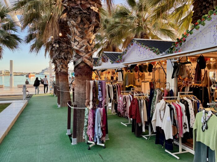 A vibrant outdoor Christmas Market featuring clothing racks under palm trees, with shoppers strolling by along a waterfront pathway. The scene captures a blend of fashion, palm trees, and a sunny atmosphere.