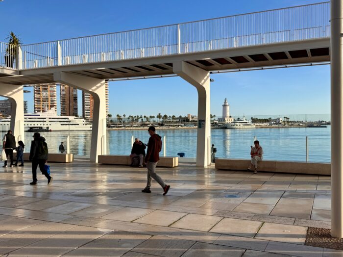 Malaga Marina waterfront promenade with people walking, sitting, and enjoying the view of a lighthouse and yachts across the water, under a blue sky. The area features sleek architecture and is bustling with visitors.