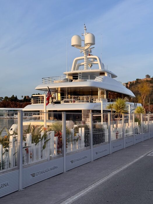 A luxurious white yacht docked at a Malaga Marina, surrounded by a modern fence and palm trees, with a scenic landscape in the background. The setting sun casts a warm glow, highlighting the yacht's sleek design.