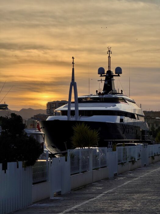 A luxurious yacht docked along a waterfront promenade during sunset, with warm golden hues in the sky and silhouettes of distant hills. Lush greenery surrounds the yacht, enhancing the serene atmosphere.