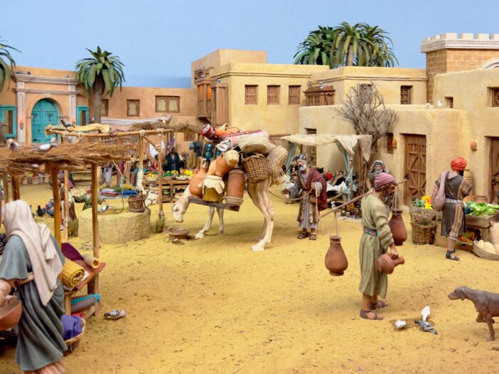Part of the Bel&eacute;n - A vibrant market scene in a desert setting, featuring figures with traditional attire, a camel carrying goods, market stalls filled with produce, and palm trees, evoking a lively atmosphere of trade and culture in an ancient town.