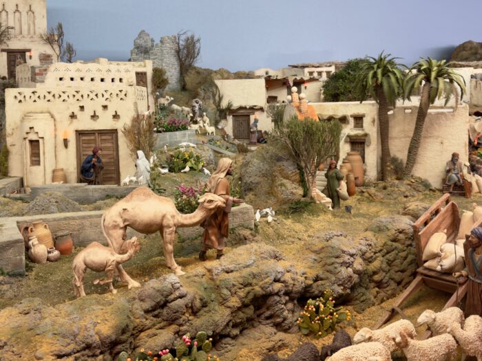 Part of the Bel&eacute;n -  A detailed diorama depicting a desert scene, featuring a camel with its calf, figures in traditional attire, palm trees, and a small village with rustic buildings, pottery, and greenery, capturing a slice of life in a historical setting.