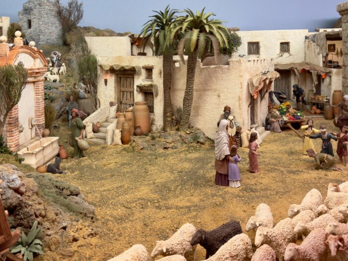 Part of the Bel&eacute;n -  A detailed model of a historical village scene featuring people interacting in a marketplace, children playing, sheep grazing, and various structures, including homes and palm trees, depicting daily life in a rural setting.