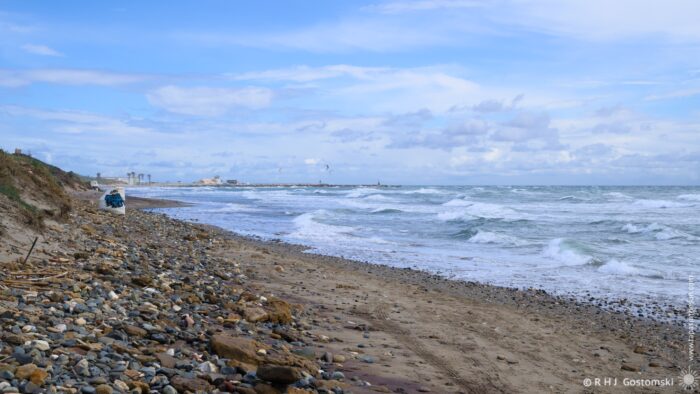 A pebble/sandy beach with waves crashing against the shore under a cloudy sky. In the distance, a pier and structures are visible along the coastline.